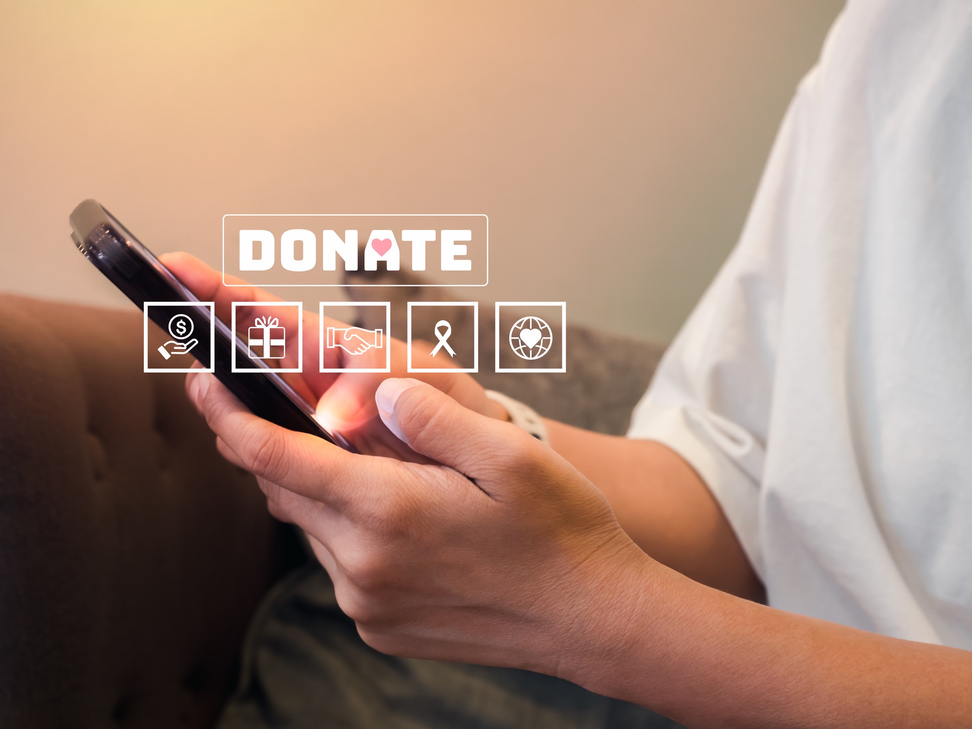 Donation online concept. Text Donate with charity and fundraising icons appear while female person in casual white shirt holding and using smart mobile phone for donating money online at her home.