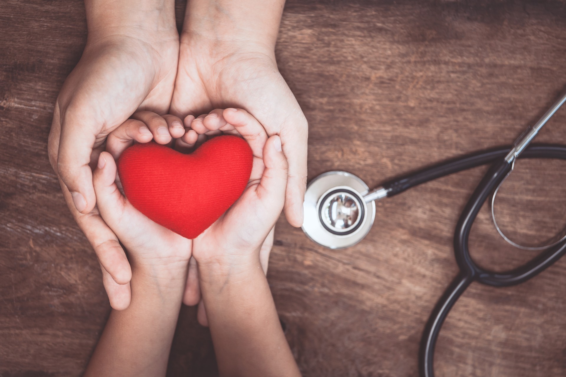 Red heart on woman and child hands and with doctor's stethoscope on wooden background